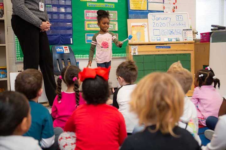 Young girl using pointer to point at her classmates while teacher watches.