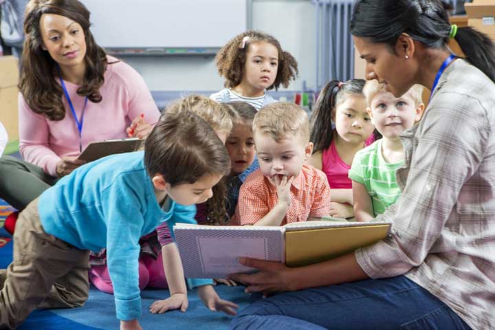 Female preschool teacher reading a story to children while an observer watches.