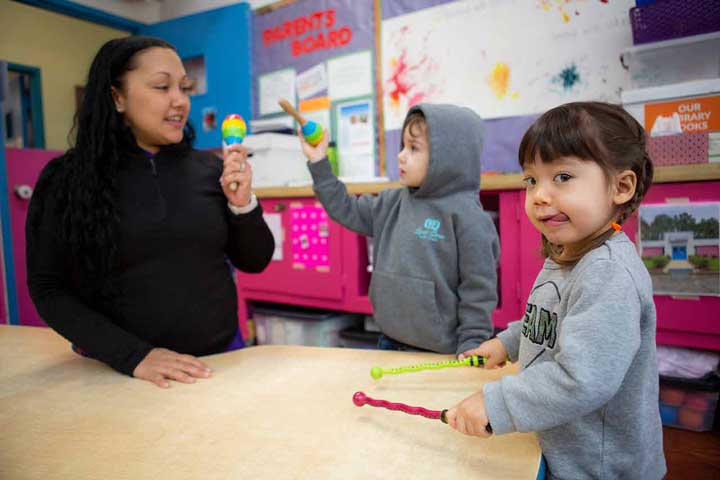 Young child playing with drum sticks while teacher holds shaker.