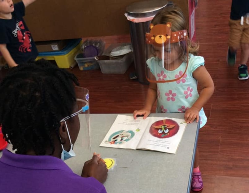 little girl wearing a face shield is reading a book and a teacher wearing a face shield is watching