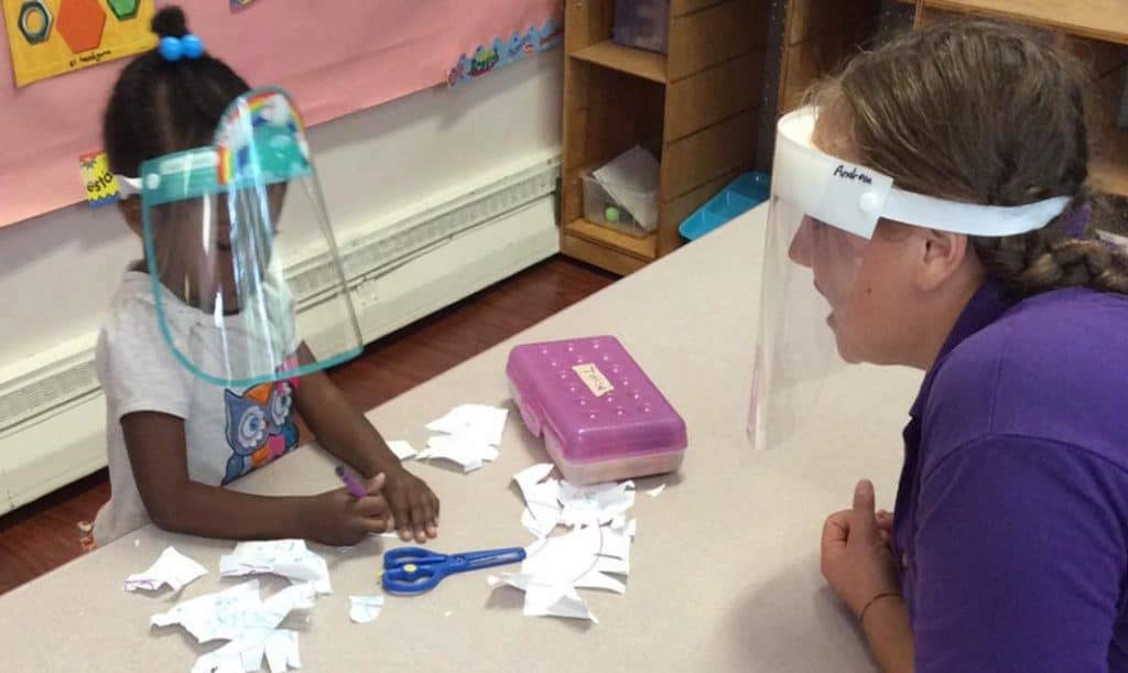little girl wearing a face shield is coloring cut up paper and her teacher (also wearing a face shield) is talking to her